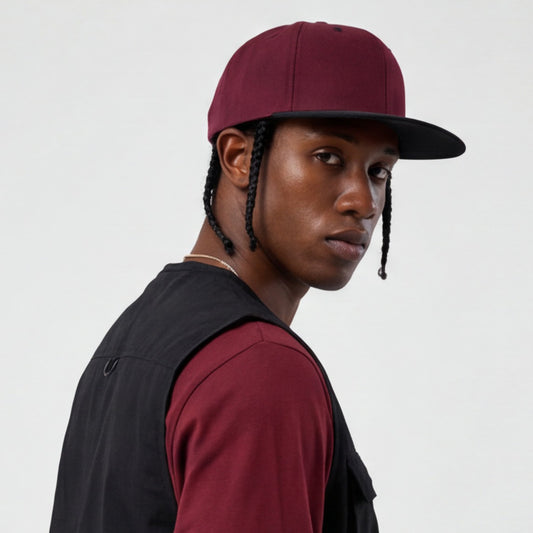 Person wearing a maroon snapback flat visor bill cap and black vest over a red shirt on a light gray background